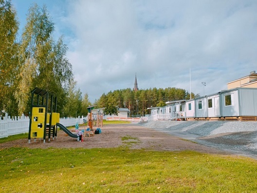 Nurmesjärven päiväkoti, julkisivukuva moduulipäiväkodista. Piha-alueella ulkoleikkivälineitä ja lapsia leikkimässä.